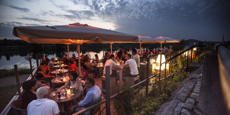 Groepen mensen zitten 's avonds op het terras aan de Maas met knusse verlichting en kaarsjes