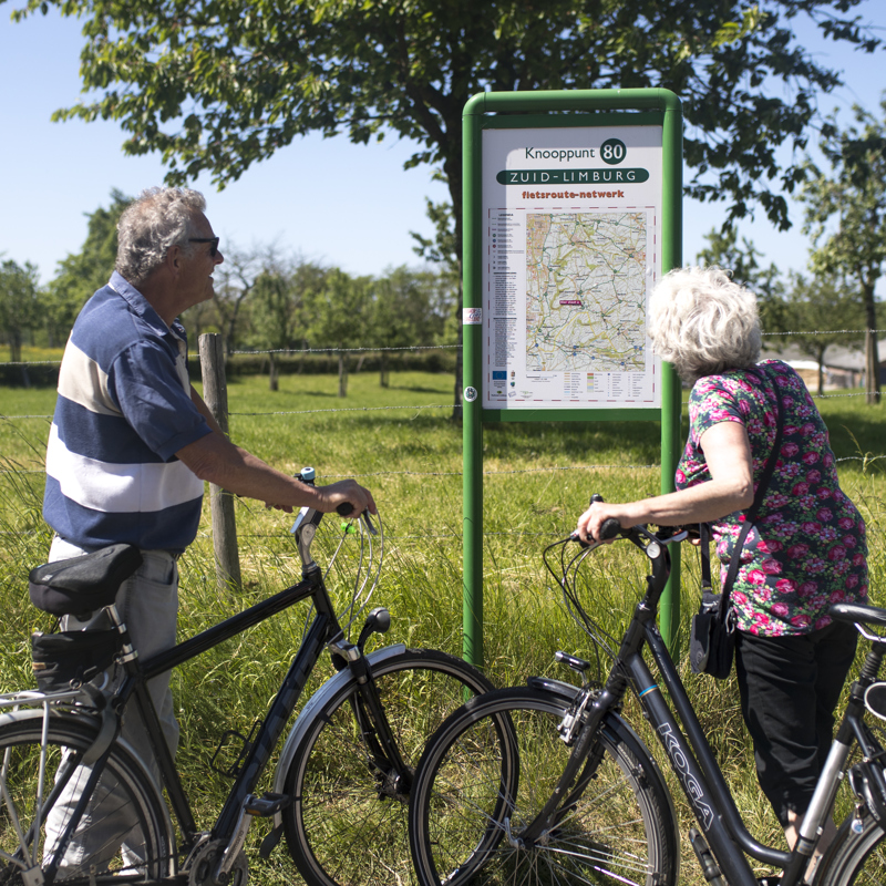 Twee ouderen mensen met fiets in hun hand kijken naar een infopaneel met fietsknooppunten